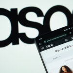 Photo of a smartphone open to the Asos website. The retailer's logo is displayed in black on a slightly off-focus white backdrop behind the phone.